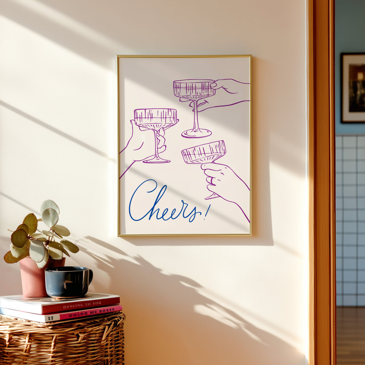 Cheers coupe glasses print shown in a gold frame above a basket of books and a pilea plant