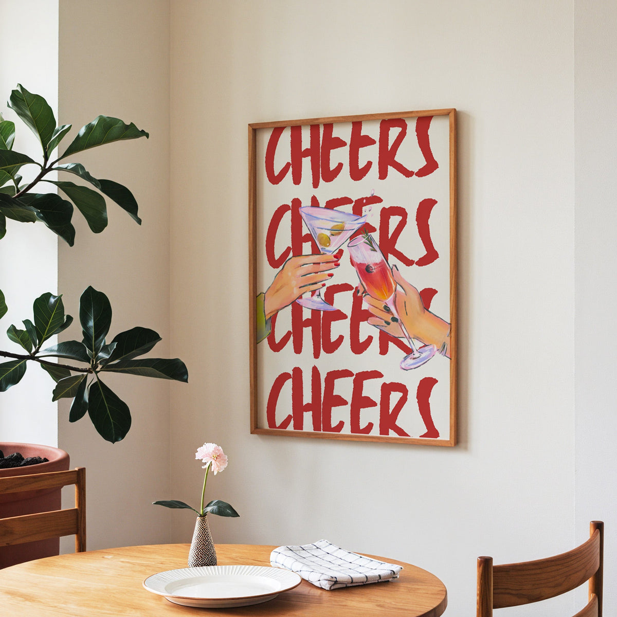 Cheers repeat type print in a walnut frame above a round table beside a fiddle-leaf fig