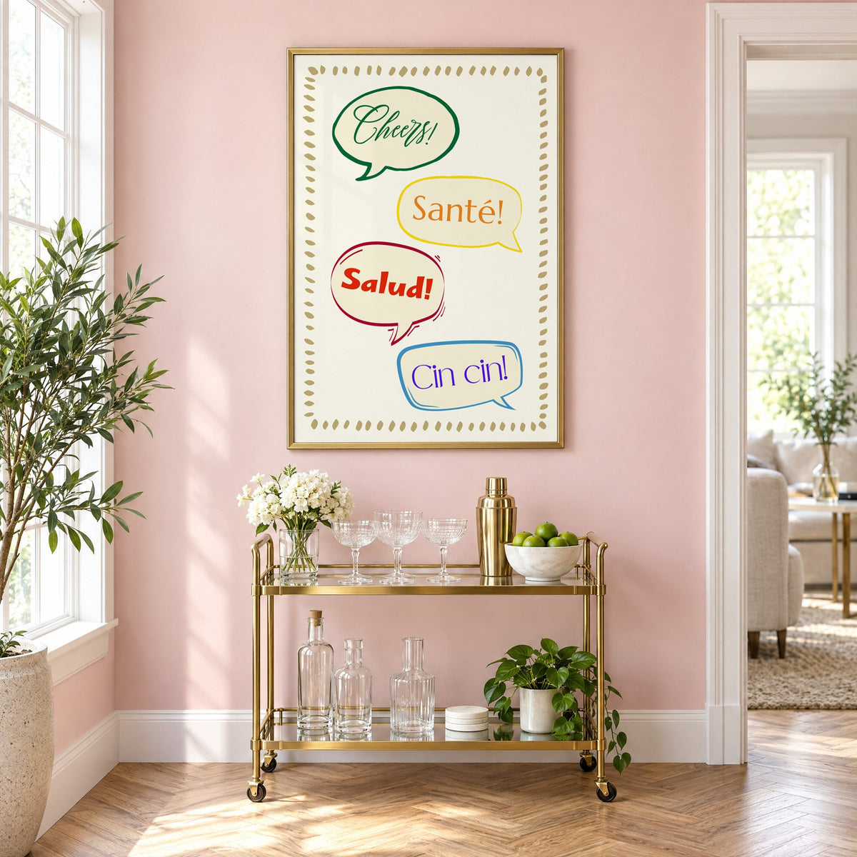 Cheers! Santé! Salud! Cin cin! speech bubbles print in a brass frame on a blush pink wall above a gold bar cart with glassware