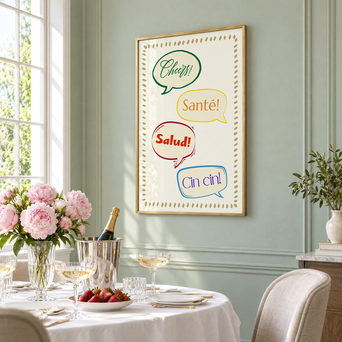 Cheers! Santé! Salud! Cin cin! speech bubbles print in a brass frame on a sage wall above a brunch table with champagne and peonies, seen at an angle