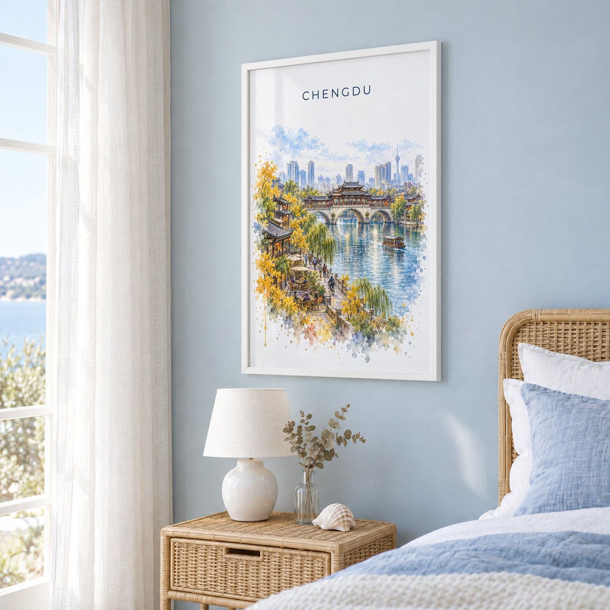 Chengdu watercolor print in a white frame on a pale blue wall beside a rattan bed in a coastal bedroom