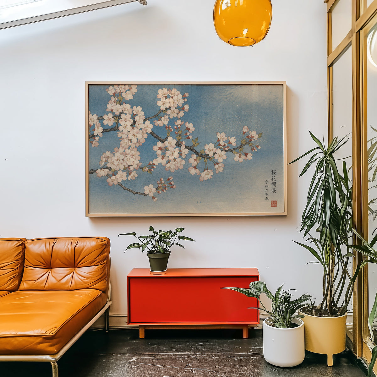 Cherry blossom branch on a blue sky print in a light wood frame above a red cabinet beside a tan leather daybed