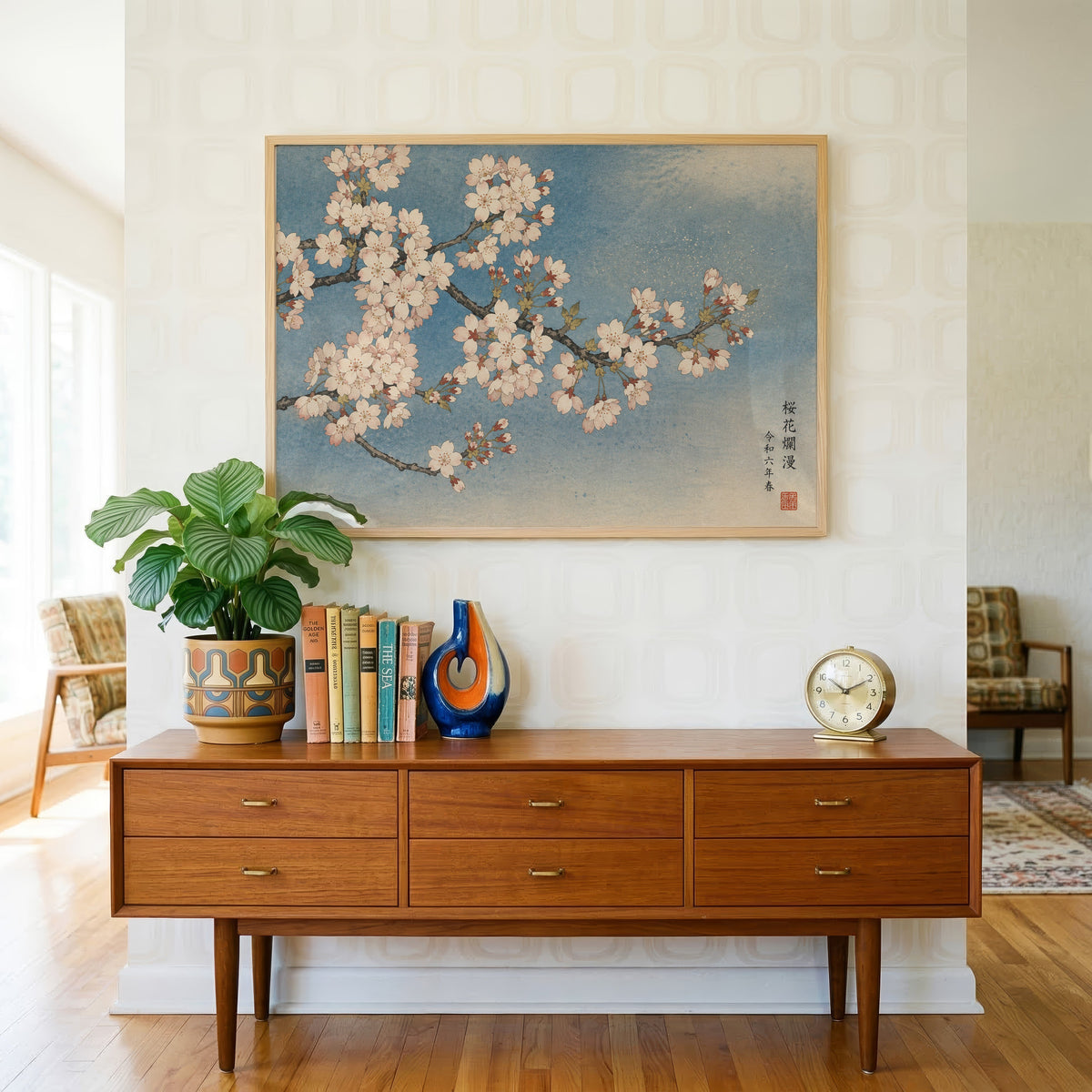 Cherry blossom branch on a blue sky print in a light oak frame above a teak credenza with books and a brass clock