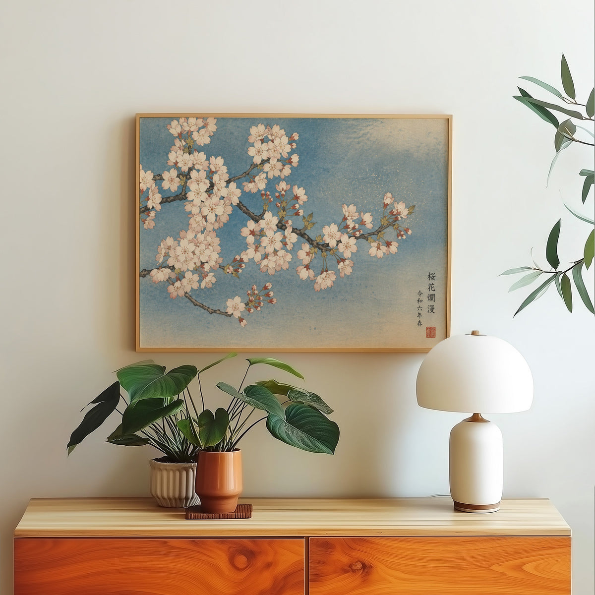 Cherry blossom branch on a blue sky print in a light oak frame above a wood sideboard with a white dome lamp