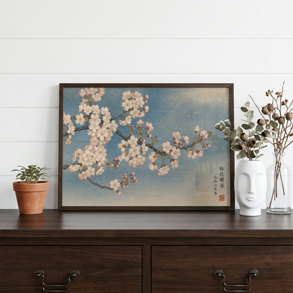 Cherry blossom branch on a blue sky print in a dark walnut frame on a dark wood dresser beside a white face vase