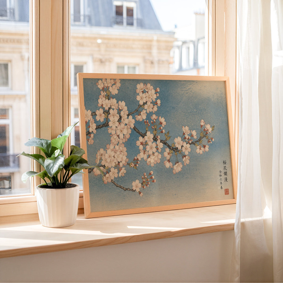 Cherry blossom branch on a blue sky print in a light wood frame leaning on a sunny windowsill by sheer curtains