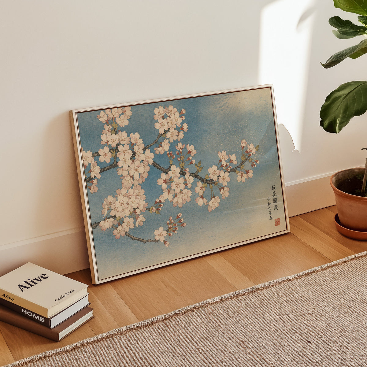 Cherry blossom branch on a blue sky print in a white floater frame leaning on a wood floor beside stacked books