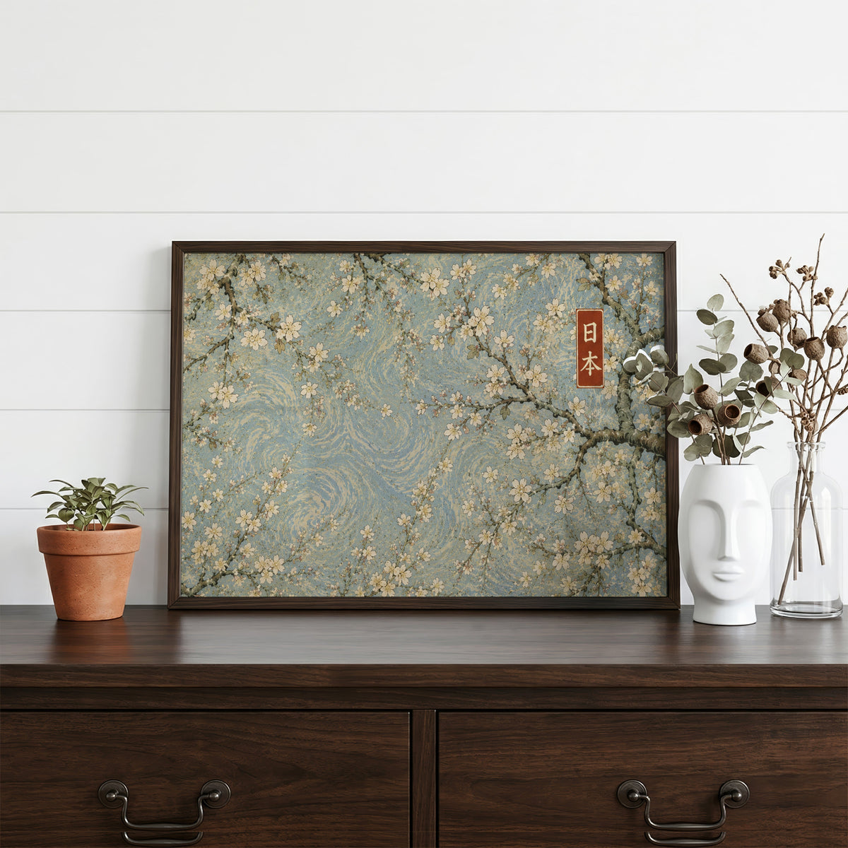 Cherry blossoms on a blue swirl sky print in a dark walnut frame on a dark wood dresser beside a white face vase