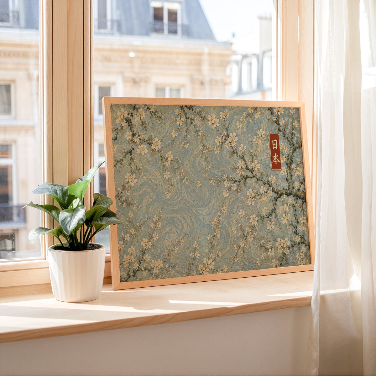 Cherry blossoms on a blue swirl sky print in a light wood frame leaning on a sunny windowsill by sheer curtains