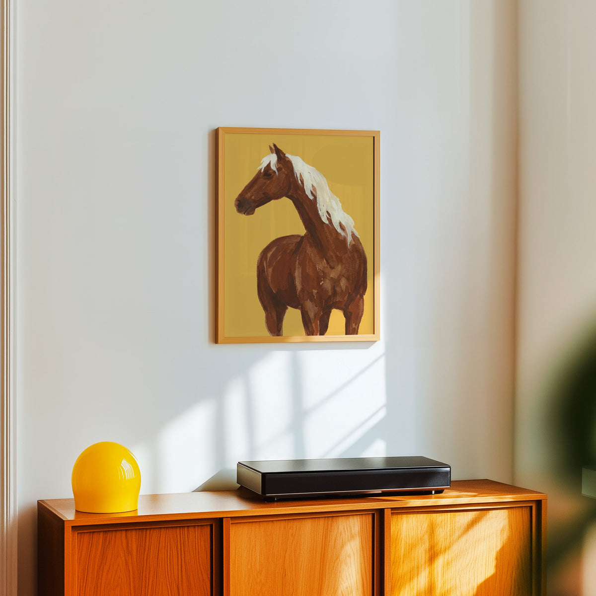 Watercolor horse portrait print in a light oak frame above a teak sideboard with a yellow lamp