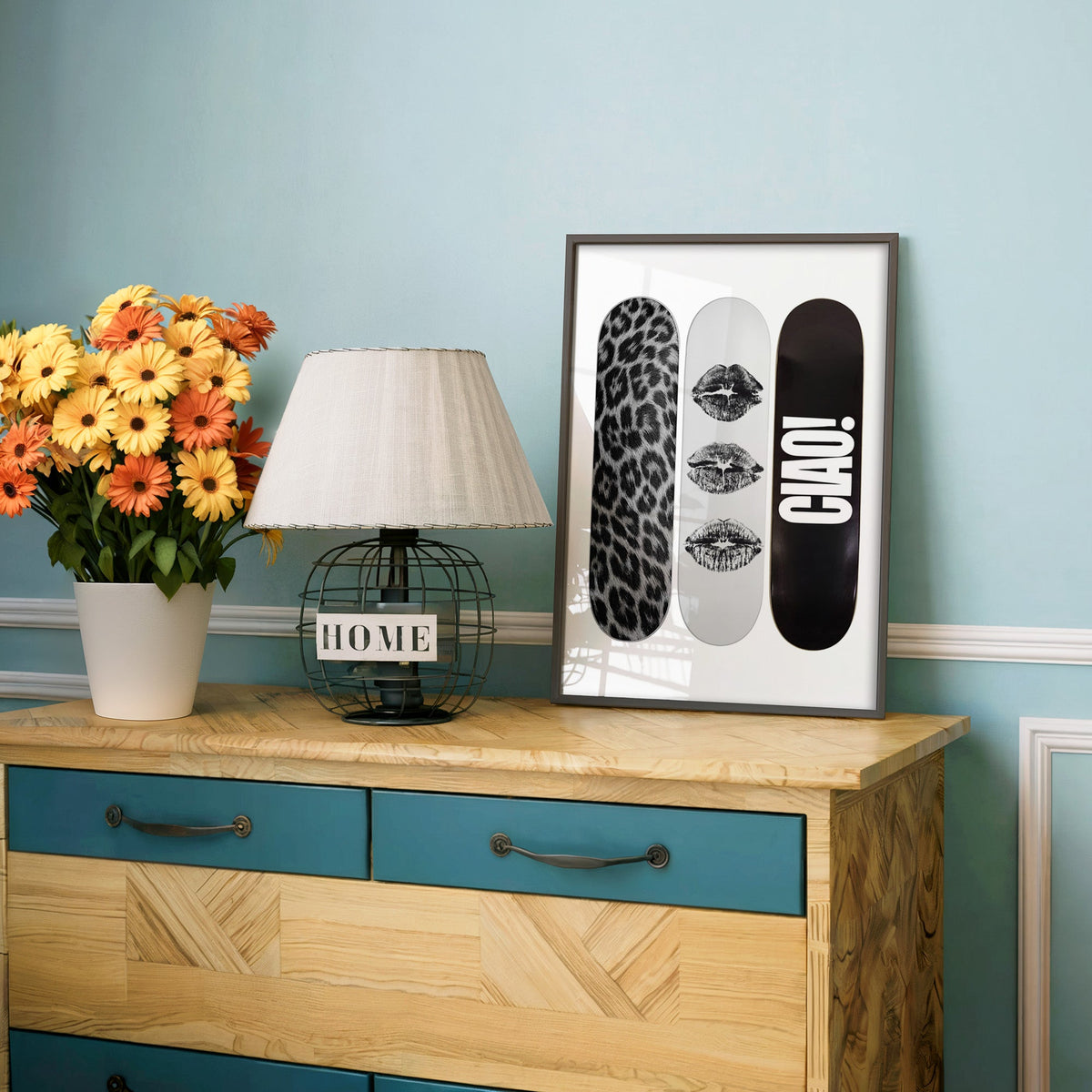 Three skateboards with fur and black lip marks print in a charcoal frame on a teal dresser beside orange daisies