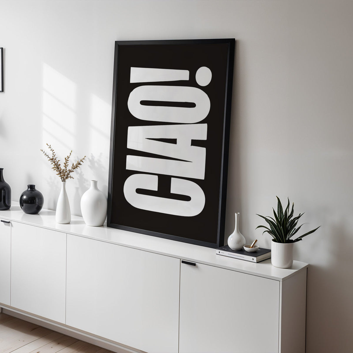 Rotated CIAO lettering on black print in a black frame on a white sideboard with black and white vases