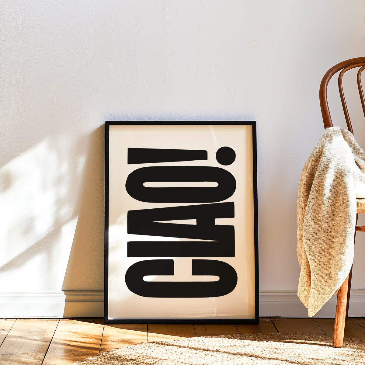 Rotated CIAO lettering on white print in a black frame leaning on a wood floor beside a bentwood chair