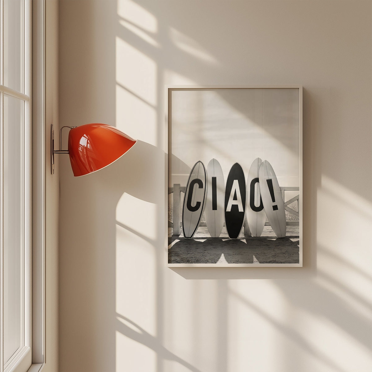 Five surfboards spelling CIAO by a beach fence print in a white frame on a sunlit wall beside a red sconce