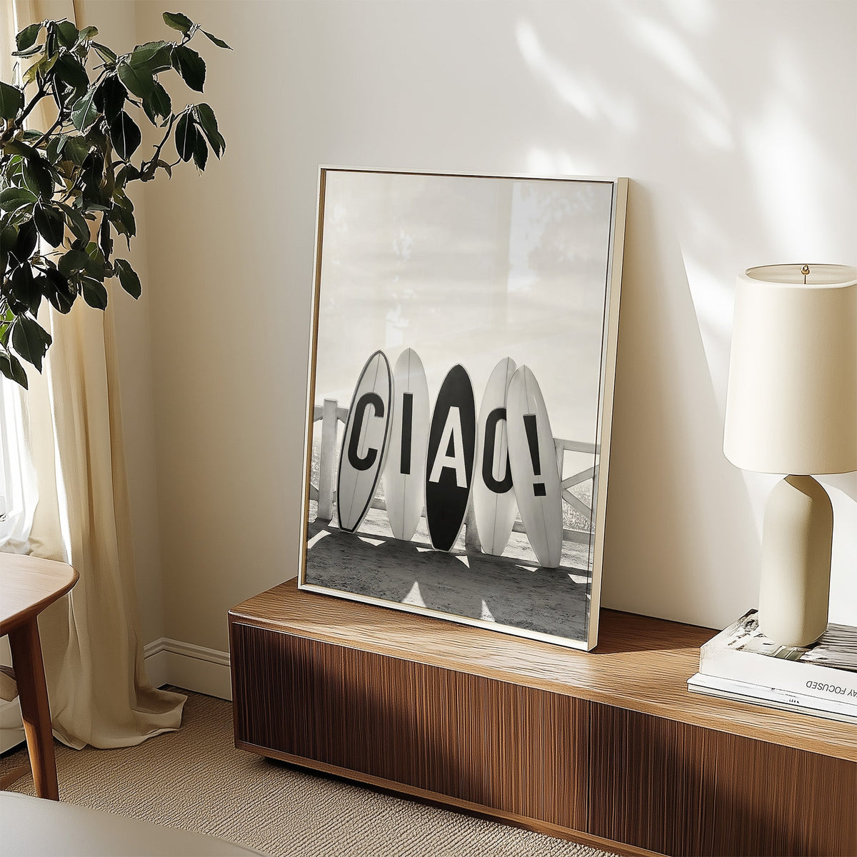 Five surfboards spelling CIAO by a beach fence print in a white frame on a walnut sideboard beside a ceramic lamp