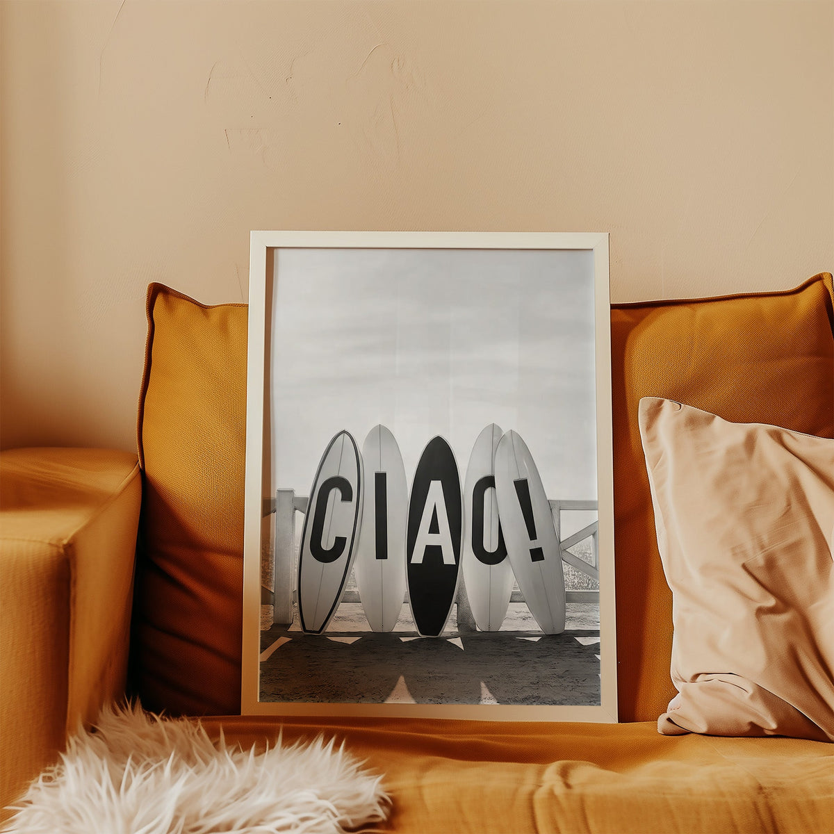 Five surfboards spelling CIAO by a beach fence print in a white frame leaning on a mustard velvet sofa