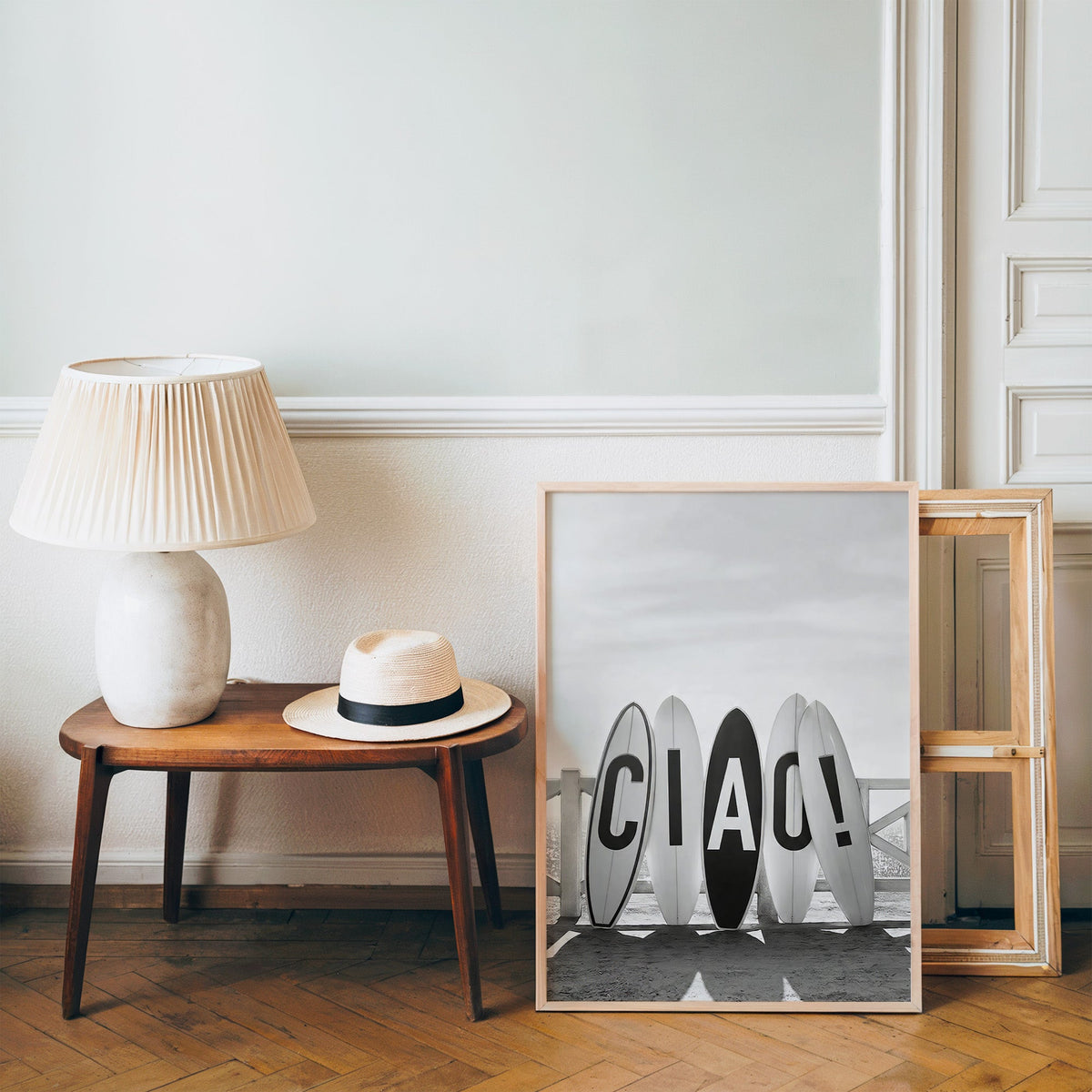 Five surfboards spelling CIAO by a beach fence print in a pine frame on a wood floor beside a lamp and straw hat