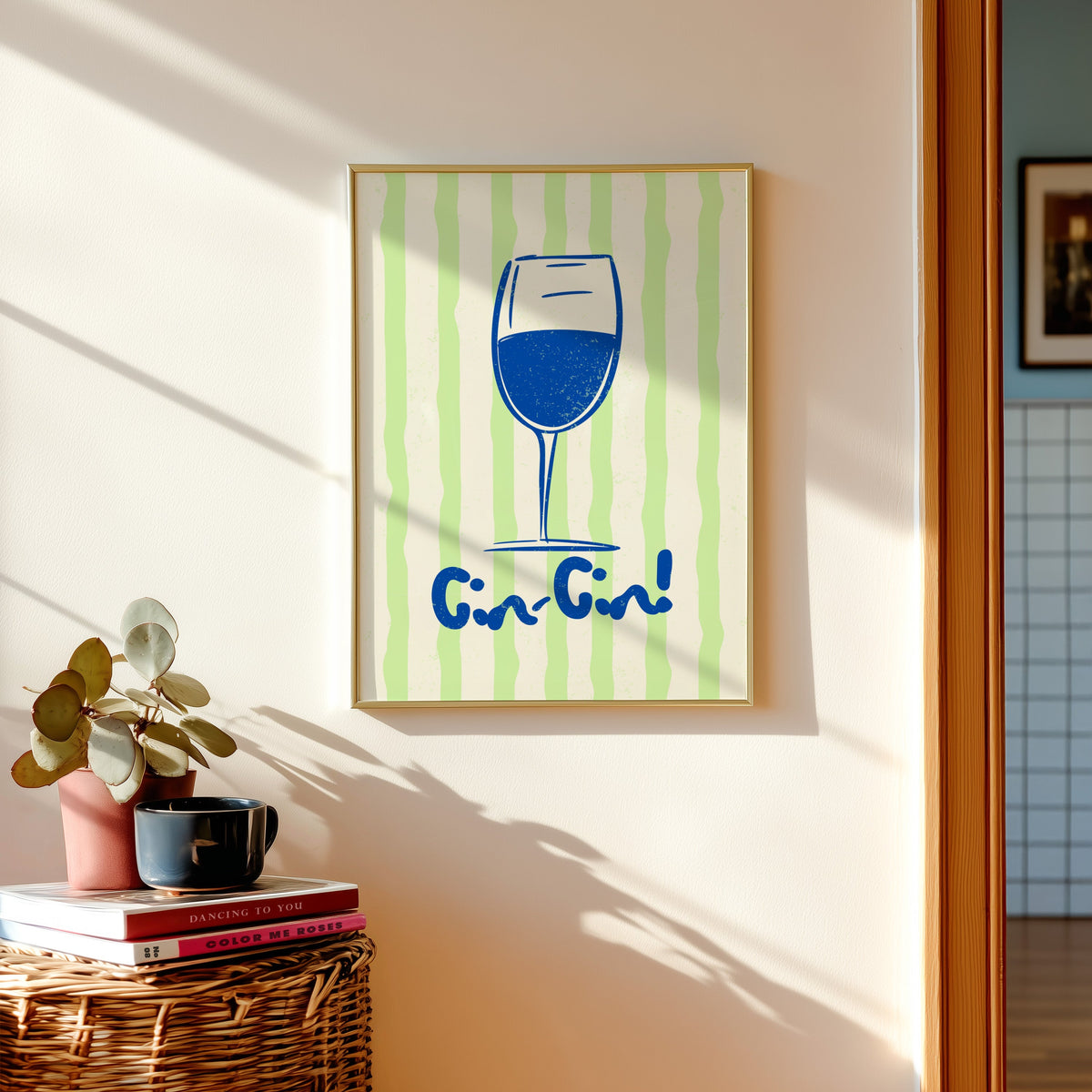 Cin Cin blue glass print shown in a gold frame above a basket of books and a pilea plant