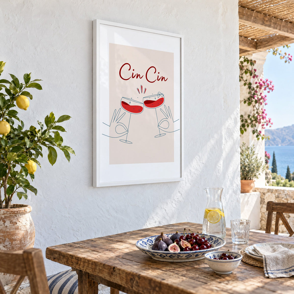Cin Cin script two hands clinking red coupes print in a white frame on a whitewashed terrace wall above a lunch table with figs and cherries, seen at an angle