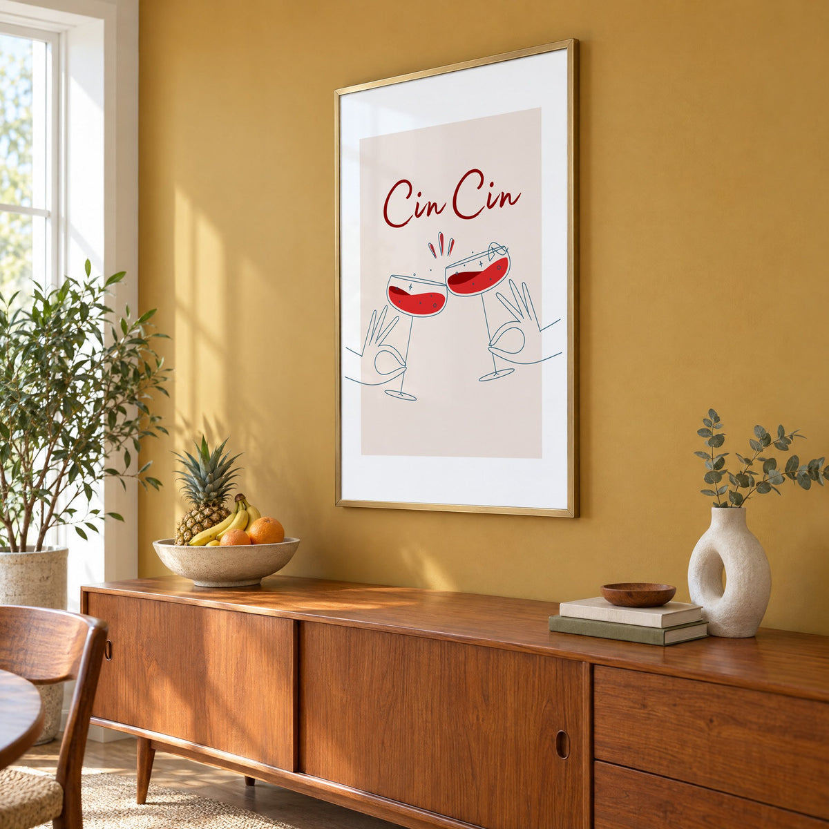 Cin Cin script two hands clinking red coupes print in a brass frame on a mustard wall above a teak sideboard with a fruit bowl, seen at an angle