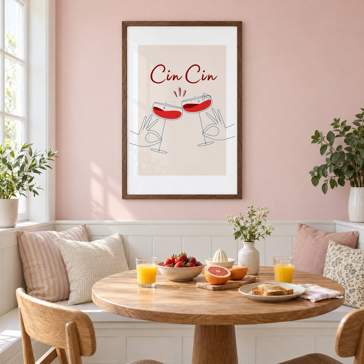 Cin Cin script two hands clinking red coupes print in a walnut frame on a pastel pink wall above a breakfast nook with strawberries and orange juice
