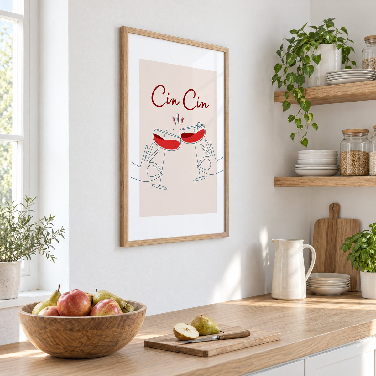 Cin Cin script two hands clinking red coupes print in an oak frame on a white kitchen wall above a counter with apples and pears, seen at an angle