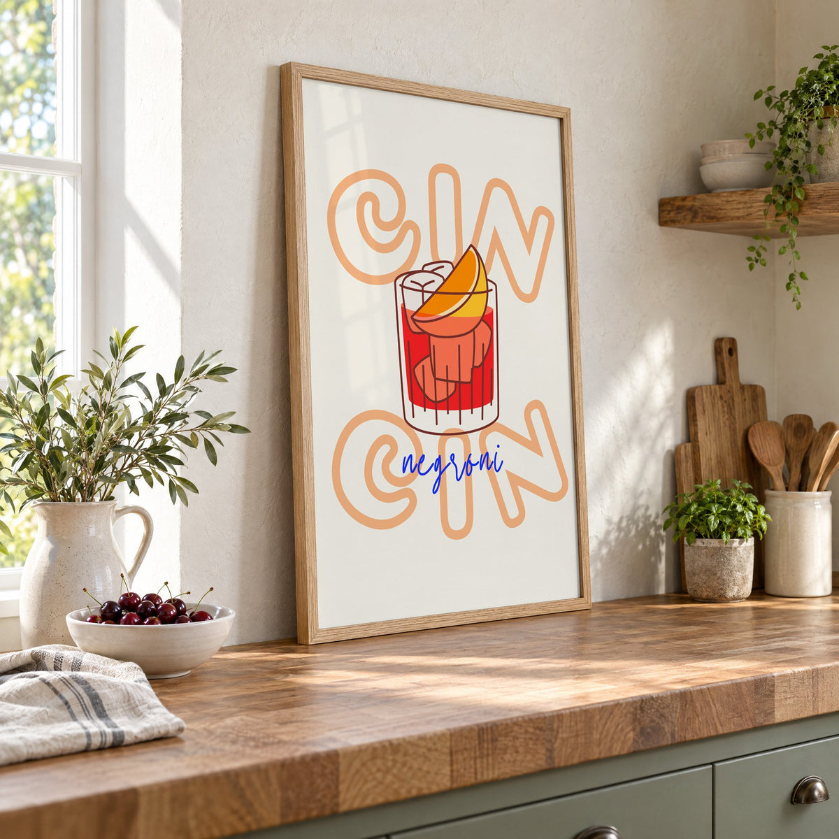 Cin Cin negroni orange slice print in an oak frame leaning on a butcher block island beside a bowl of cherries