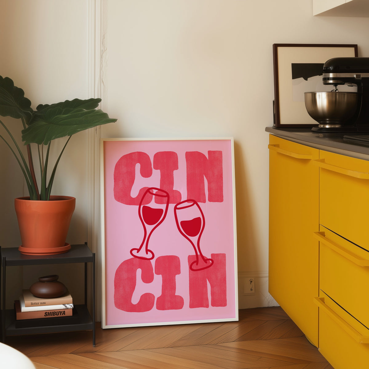 Cin Cin pink type print in a white frame leaning on parquet beside a bright yellow kitchen