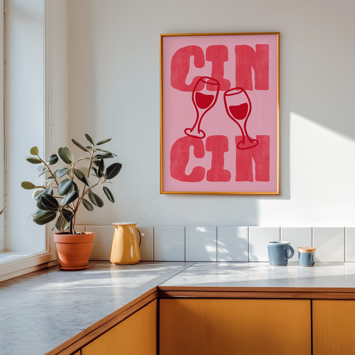 Cin Cin pink type print shown in a gold frame above a marble counter with a yellow ceramic jug