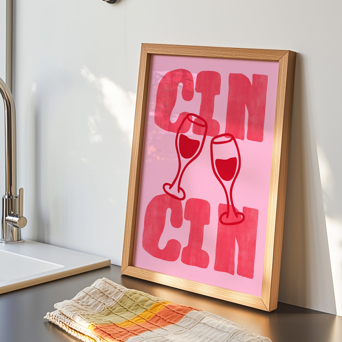Cin Cin pink type print in an oak frame by the kitchen sink beside a striped tea towel