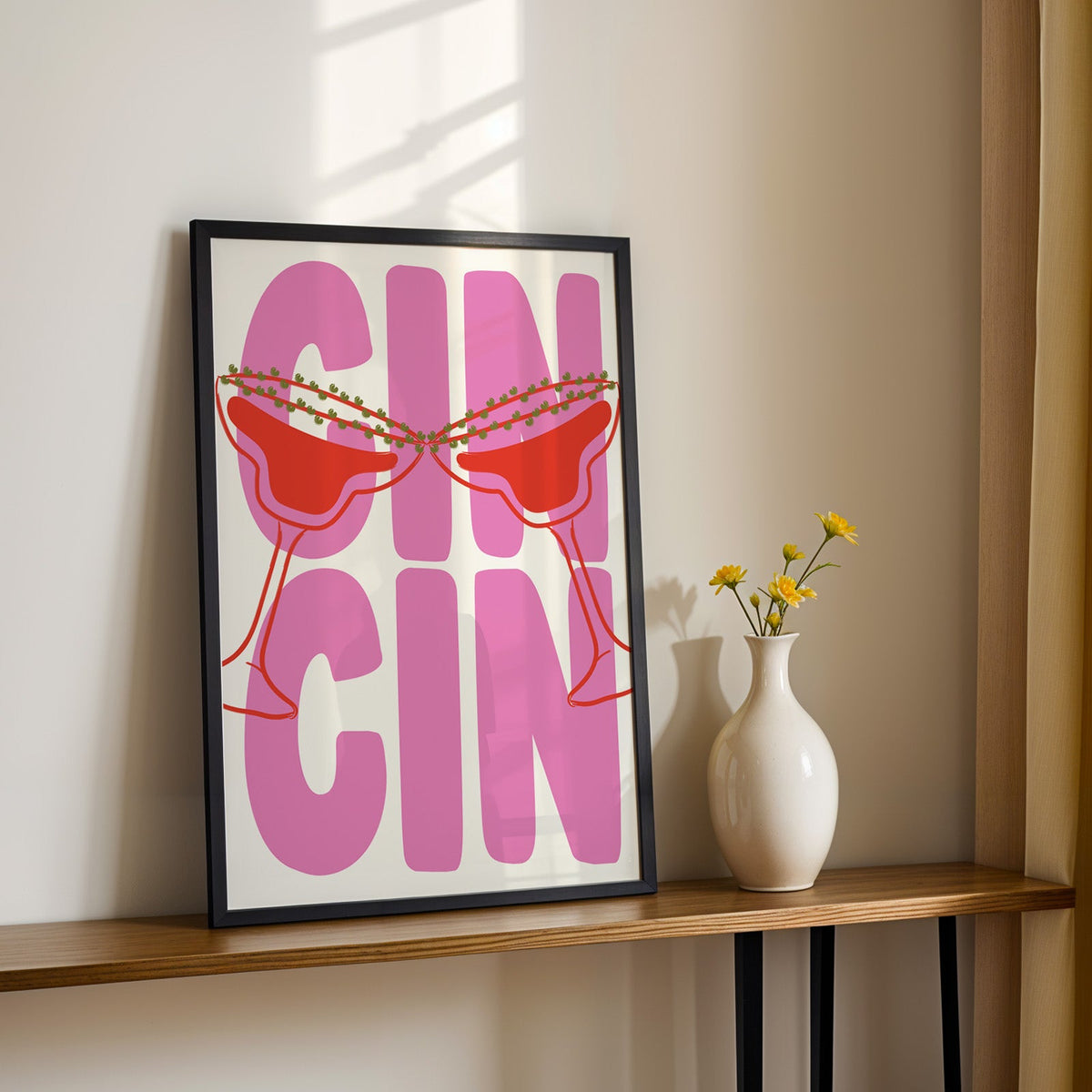 Cin cin pink coupe print in a black frame on a console with yellow daisies