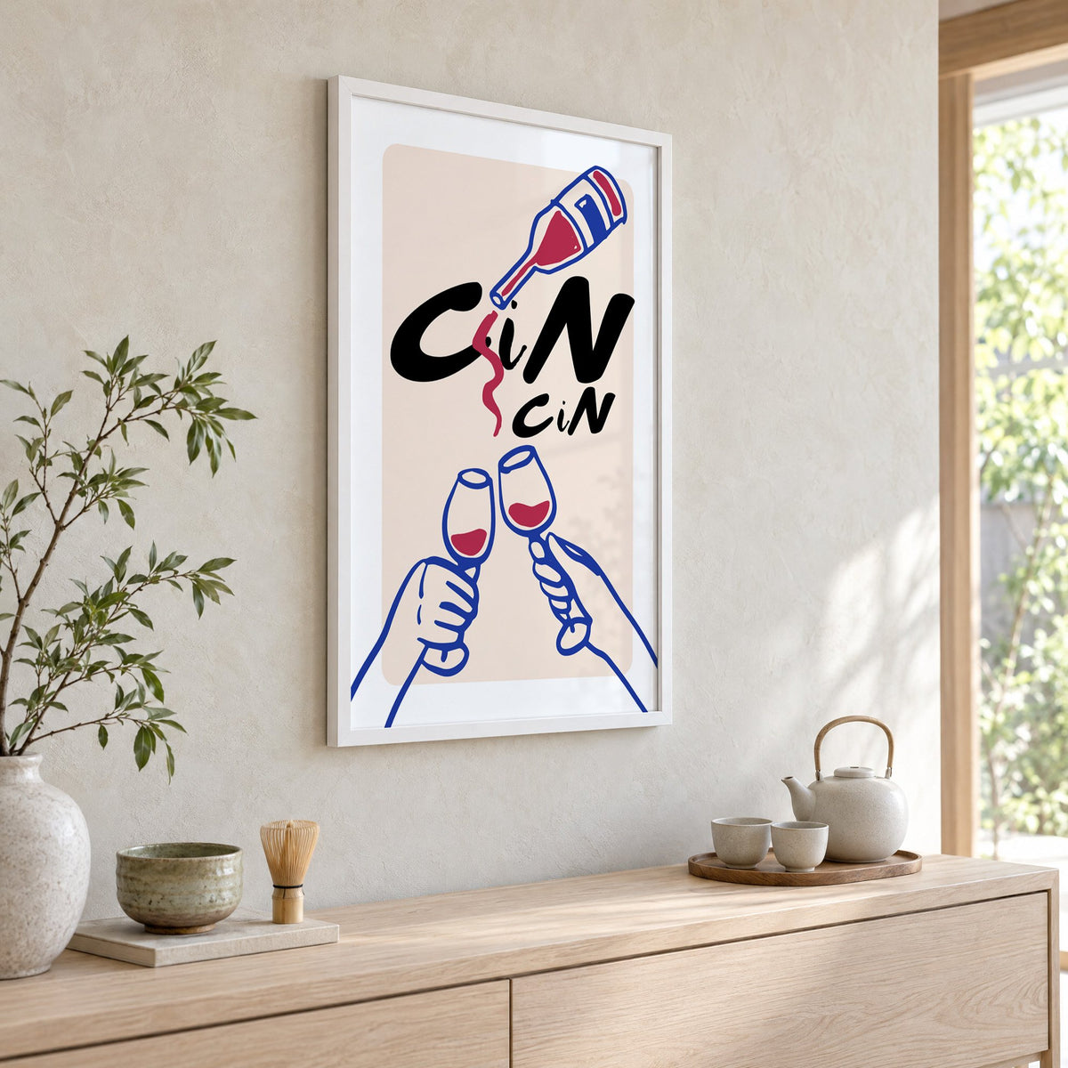 CiN CiN red wine toast print in a white frame above a pale wood console with a matcha tea set
