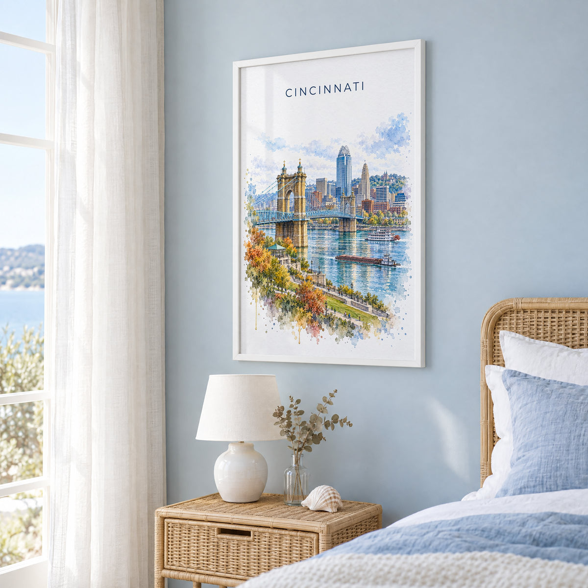 Cincinnati watercolor print in a white frame on a pale blue wall beside a rattan bed in a coastal bedroom