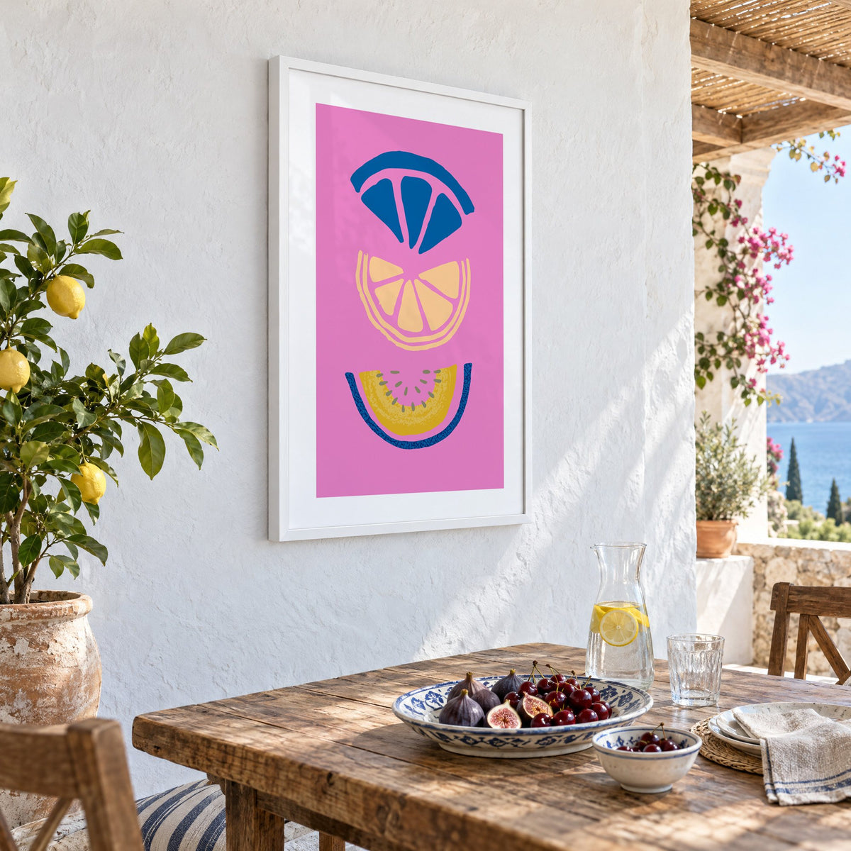 stacked citrus and melon slices hot pink print in a white frame on a whitewashed terrace wall above a lunch table with figs and cherries, seen at an angle