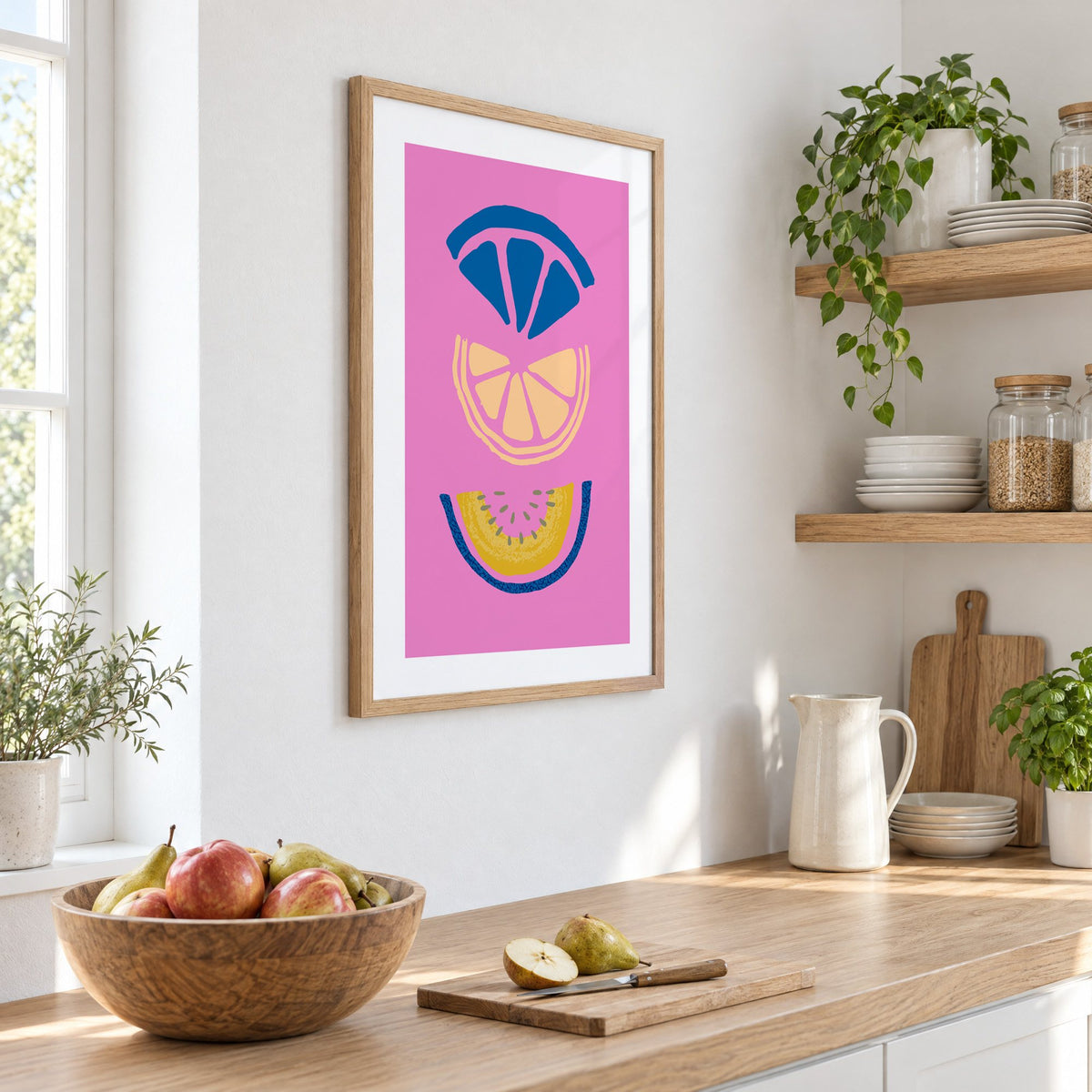 stacked citrus and melon slices hot pink print in an oak frame on a white kitchen wall above a counter with apples and pears, seen at an angle