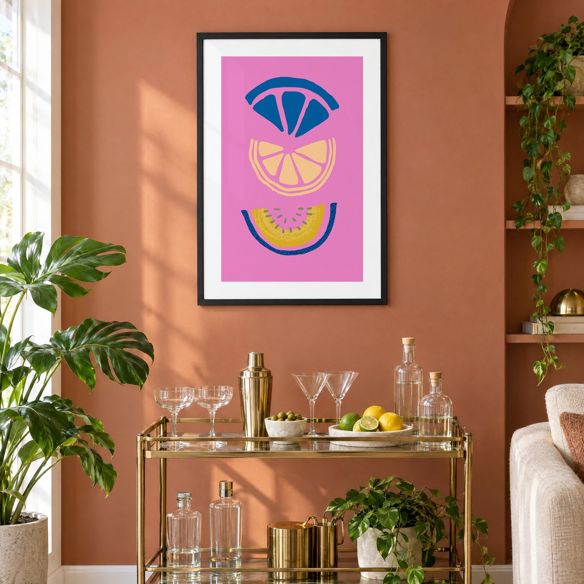 stacked citrus and melon slices hot pink print in a black frame on a terracotta wall above a brass bar cart with coupes and citrus
