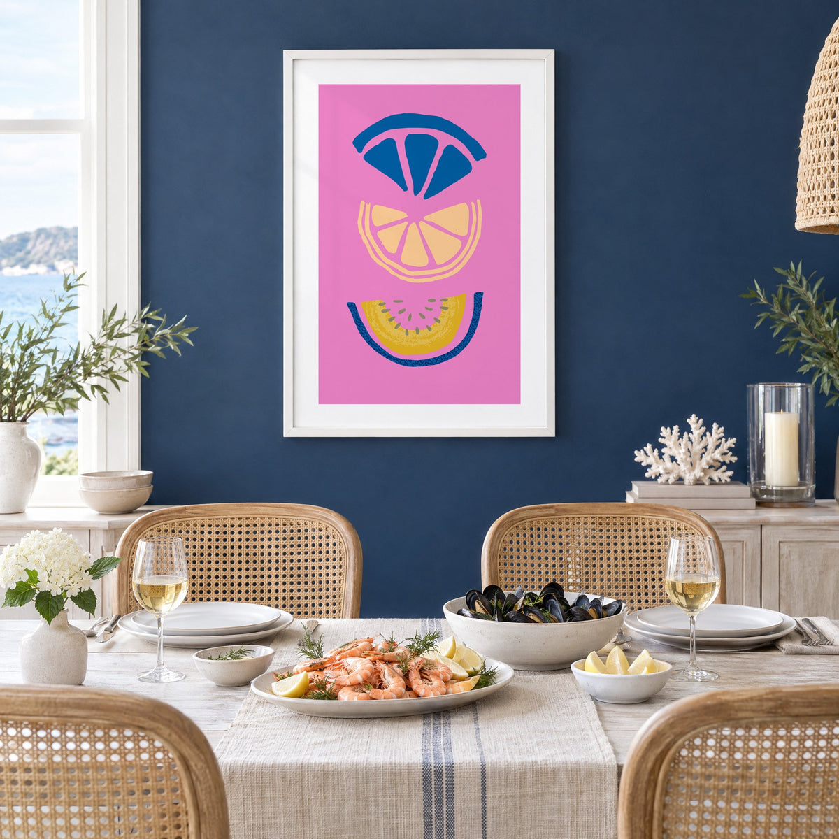 stacked citrus and melon slices hot pink print in a white frame on a navy wall above a dining table set with shrimp and mussels