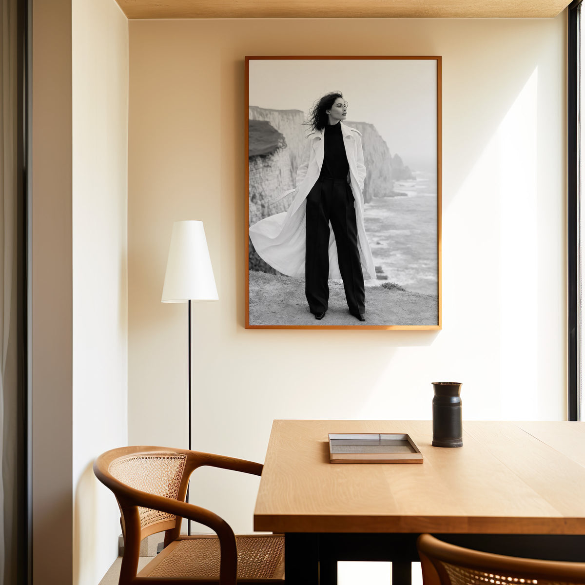 Black and white model in white trench on cliffs print in an oak frame above a dining table and cane chair