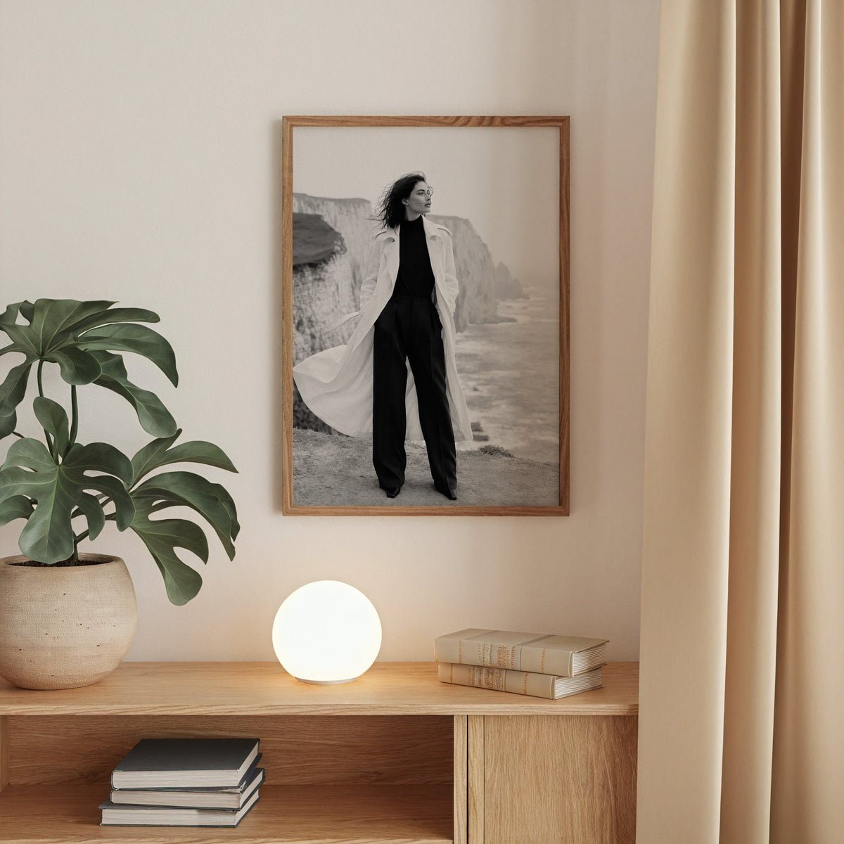 Black and white model in white trench on cliffs print in a walnut frame above an oak cabinet with a glowing globe lamp