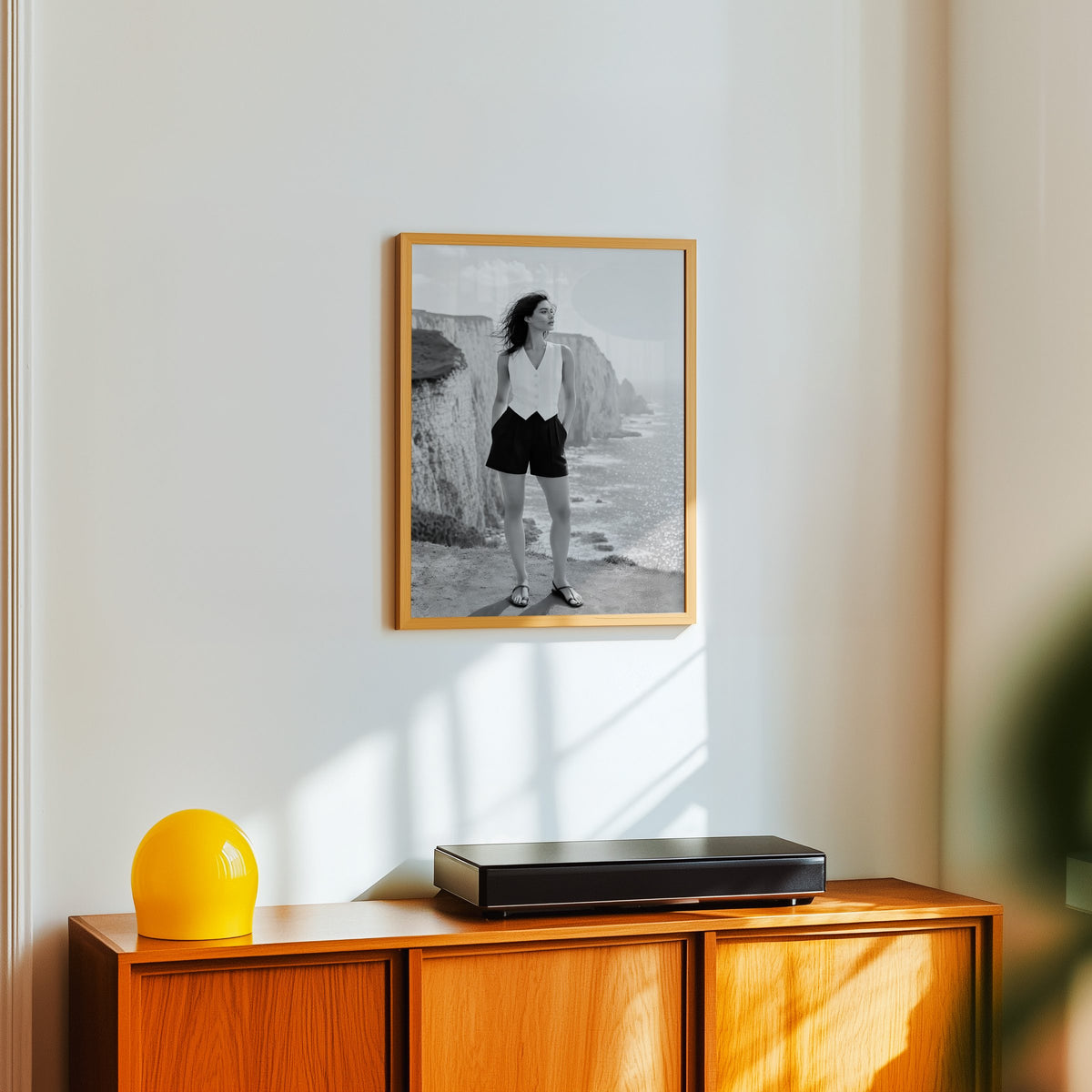 Black and white model in white vest on cliffs print in an oak frame above a teak sideboard with a yellow glass lamp