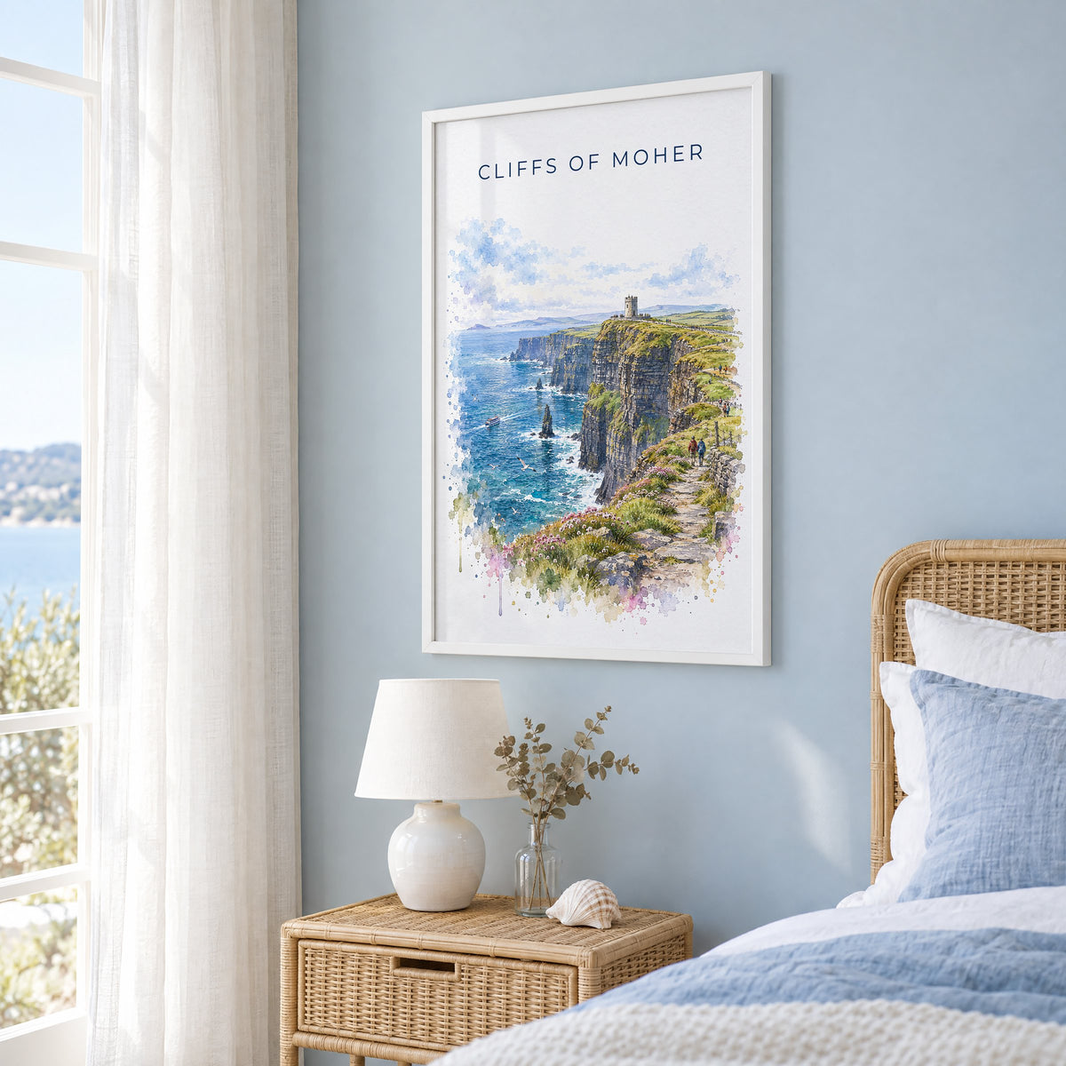 Cliffs of Moher watercolor print in a white frame on a pale blue wall beside a rattan bed in a coastal bedroom