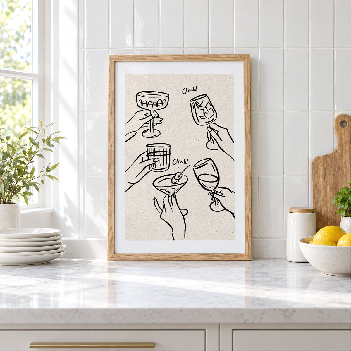 Clink! black line-art cocktail toast print in an oak frame leaning on a marble counter by white tiles and lemons