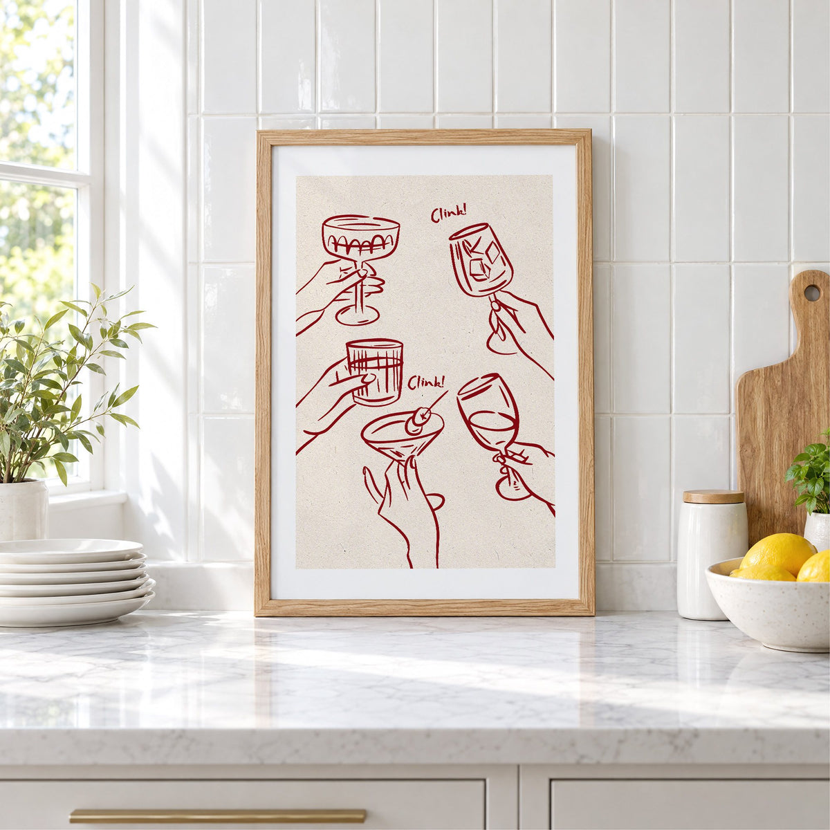 Clink! wine-red cocktail toast print in an oak frame leaning on a marble counter by white tiles and lemons