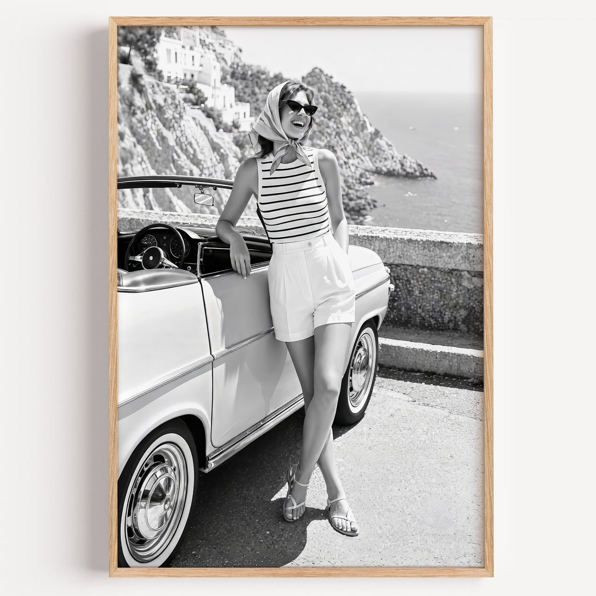 Woman leaning on vintage convertible by the sea print in a light oak frame on a plain white wall