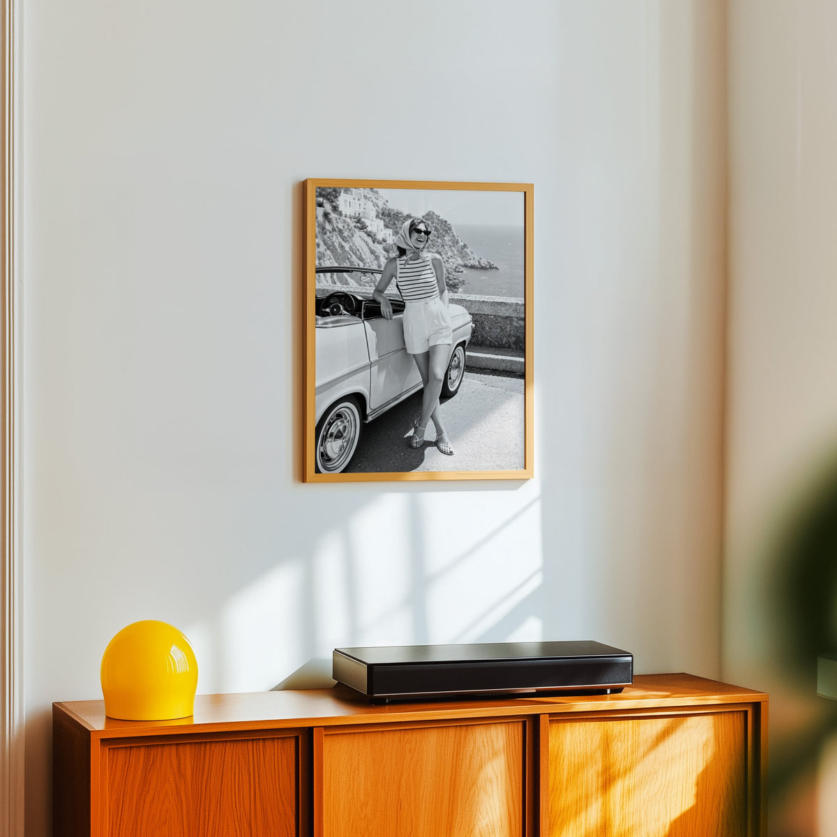 Woman leaning on vintage convertible by the sea print in an oak frame above a teak sideboard with a yellow glass lamp