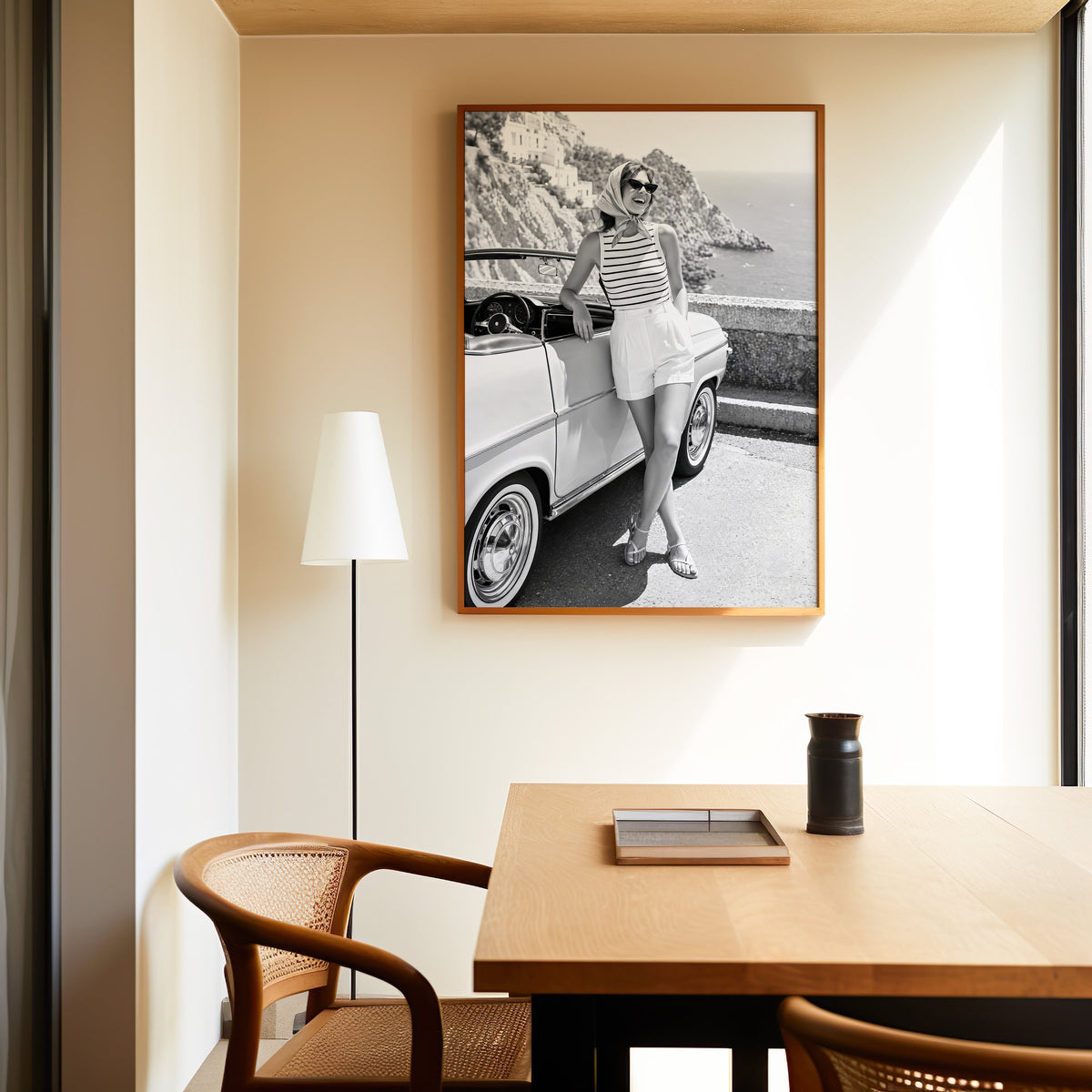 Woman leaning on vintage convertible by the sea print in an oak frame above a dining table and cane chair
