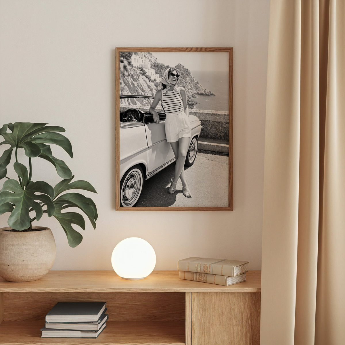 Woman leaning on vintage convertible by the sea print in a walnut frame above an oak cabinet with a glowing globe lamp