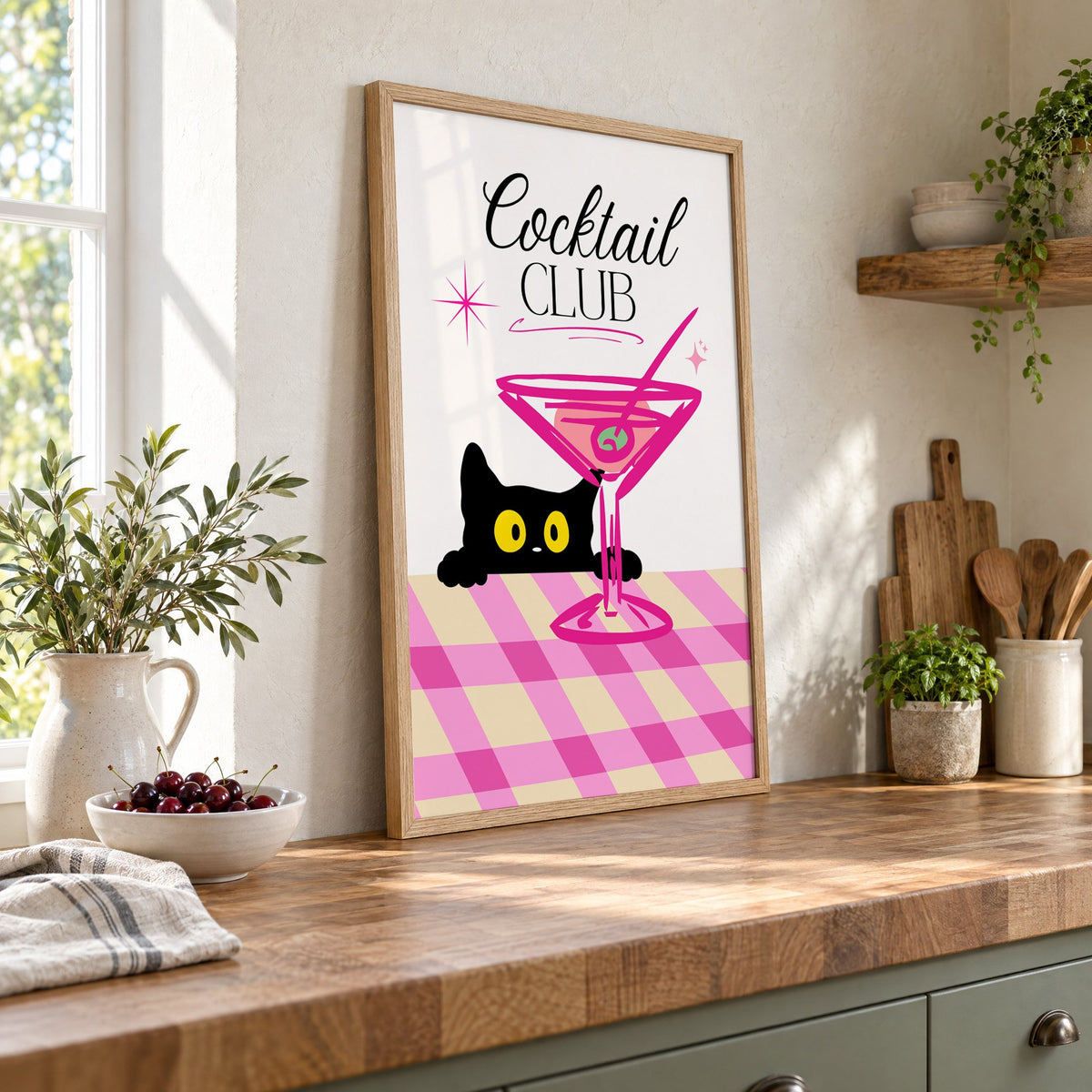 Cocktail Club black cat martini print in an oak frame leaning on a butcher block island beside a bowl of cherries