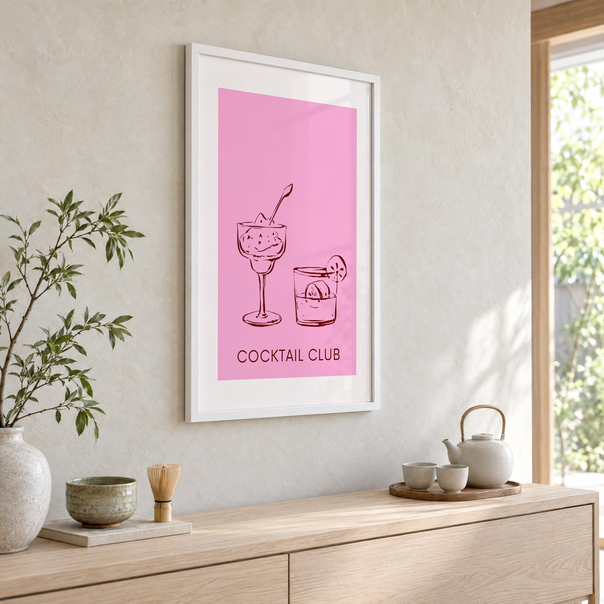 Cocktail Club sketched glasses pink print in a white frame above a pale wood console with a matcha tea set