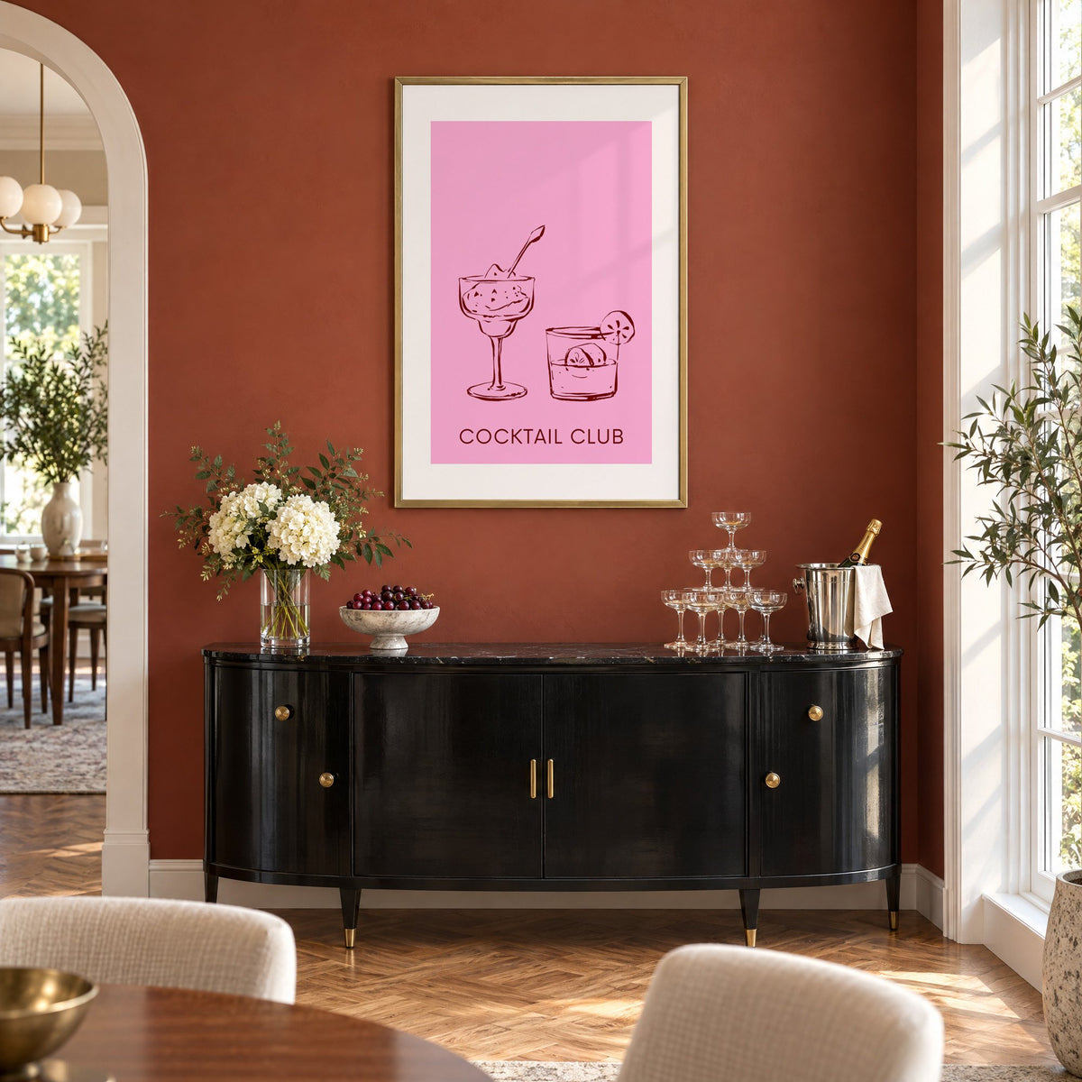 Cocktail Club sketched glasses pink print in a brass frame on a terracotta wall above a black buffet with a champagne tower